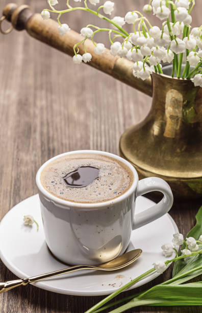 Hot coffee in white cup and spoon, copper coffee pot with white lilies of the valley, twigs convallaria with leaves on  wooden table.